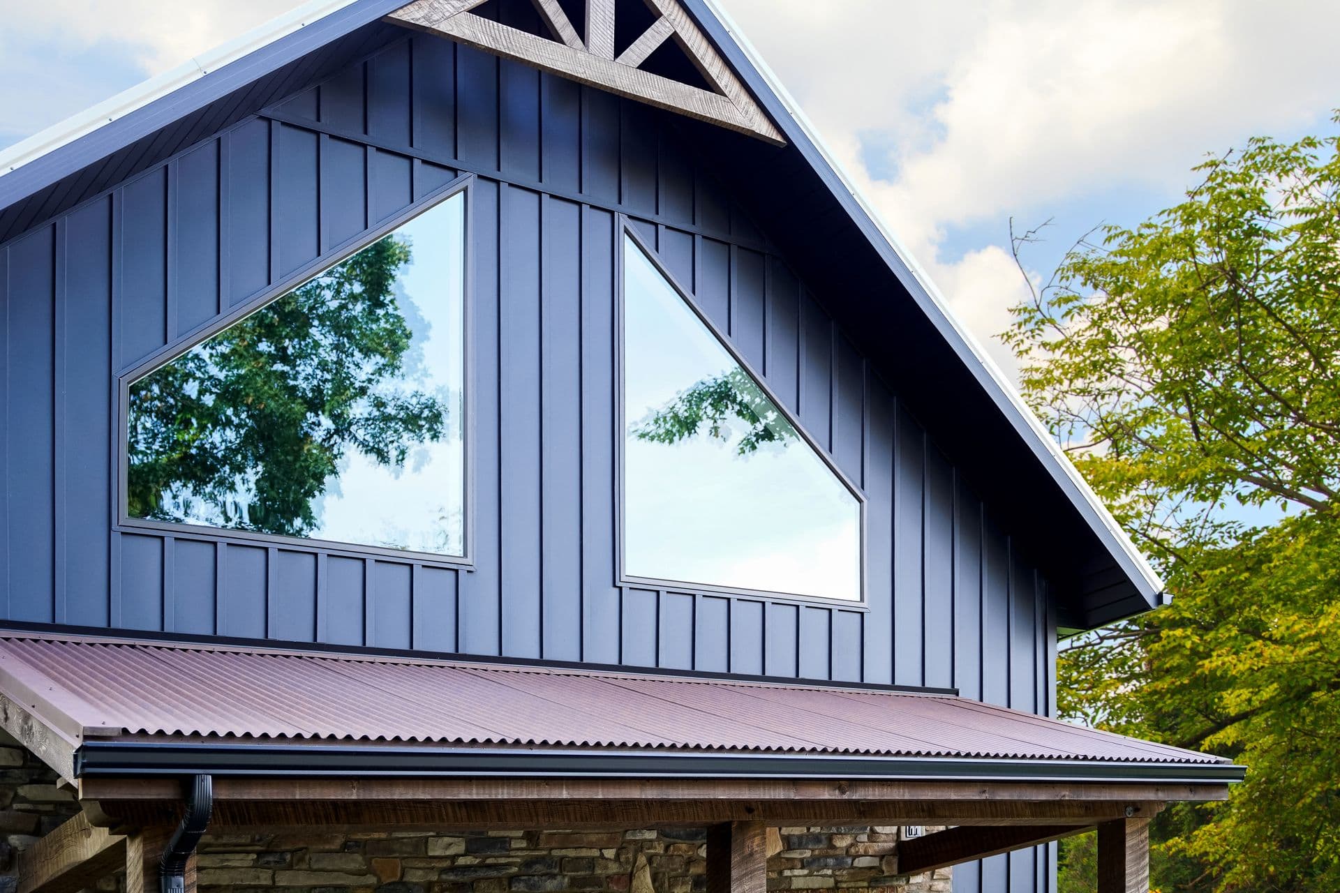 A modern cabin with dark blue siding and geometric windows reflects trees against a partly cloudy sky.