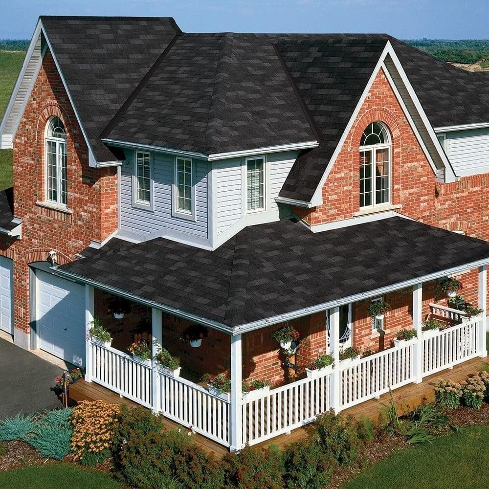 A two-story brick house with a black shingle roof and white porch railing, surrounded by landscaped gardens.