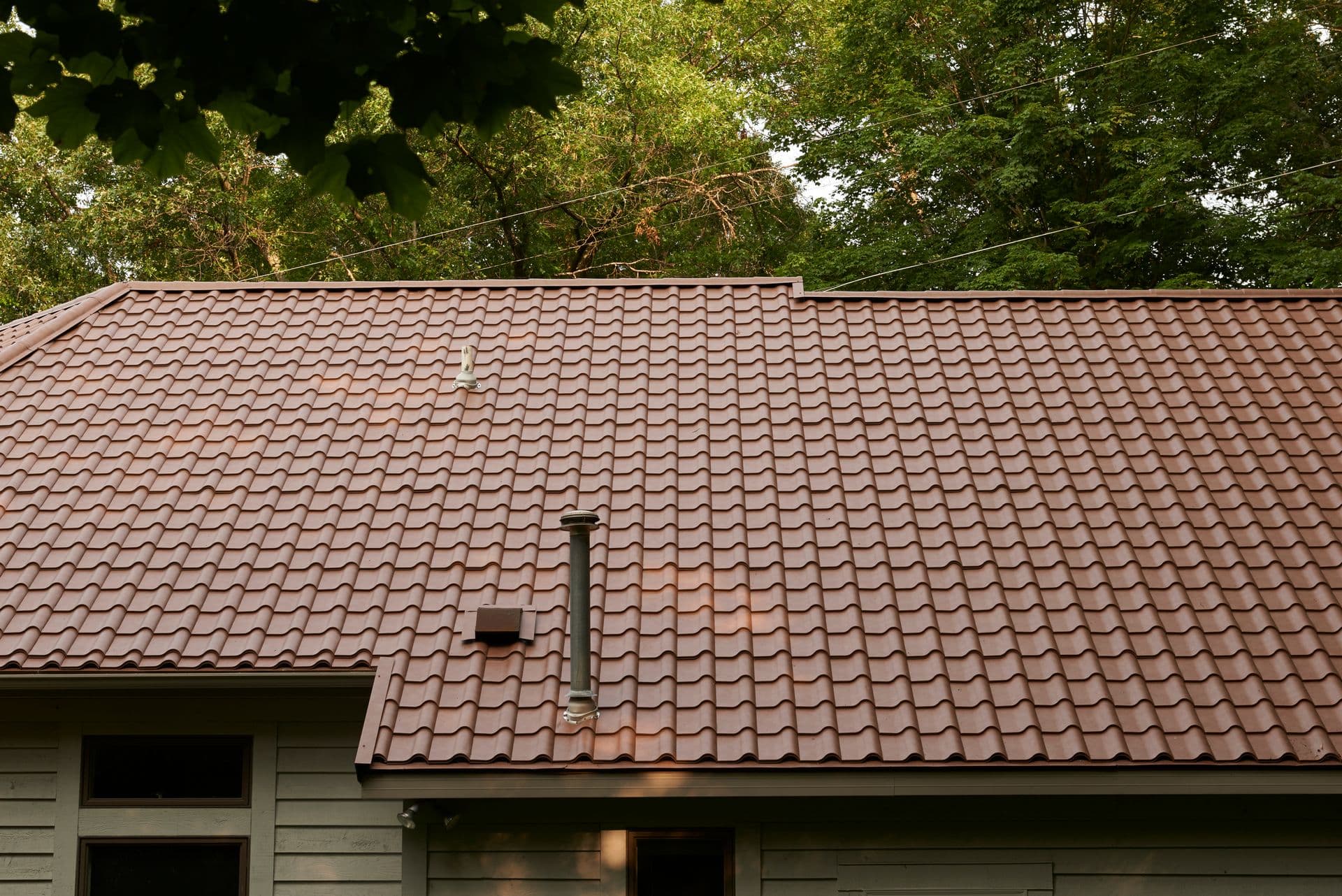 A close-up of a brown tiled roof with a chimney and surrounded by green trees.
