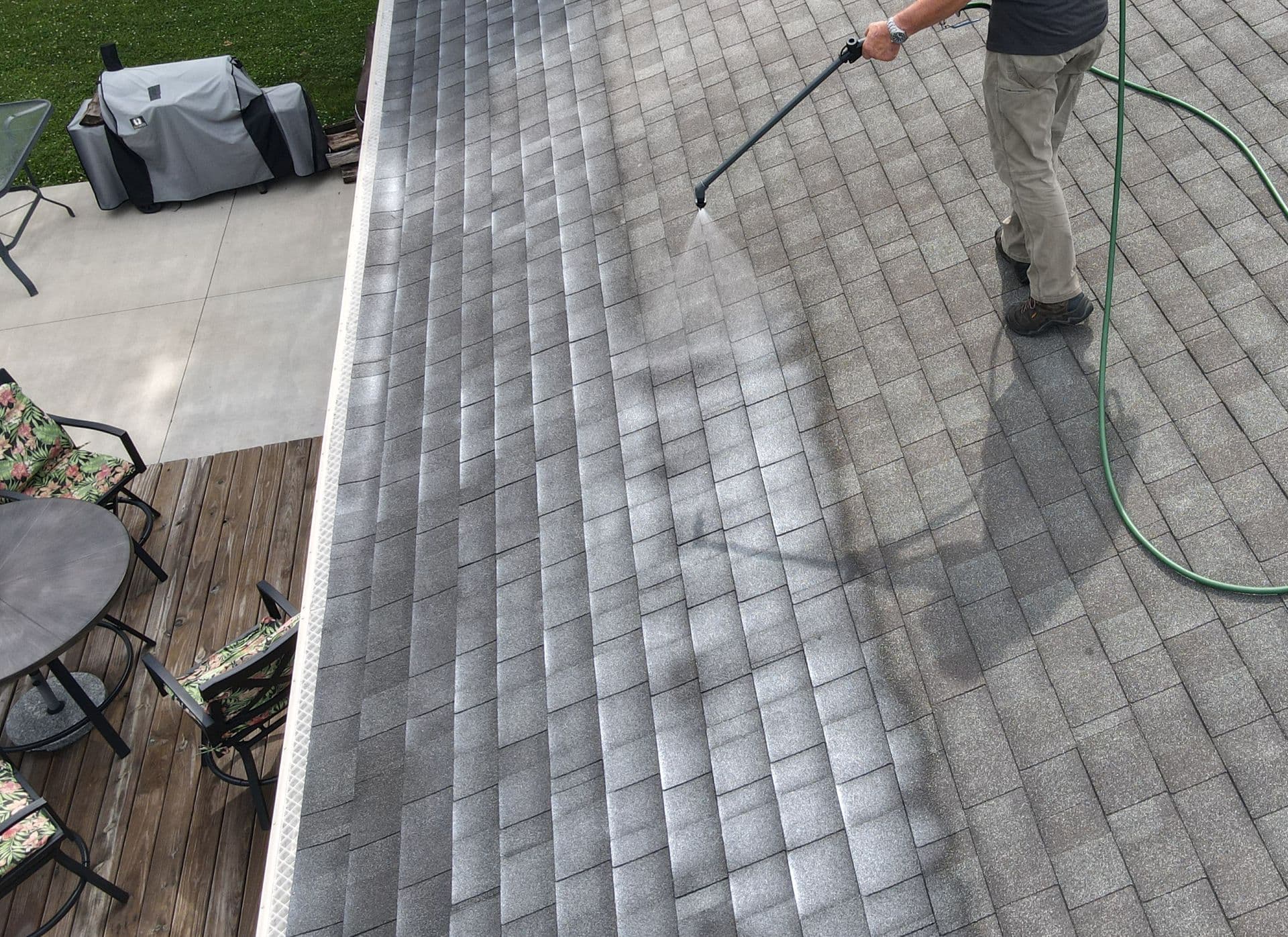 A person uses a pressure washer to clean a gray shingled roof, with a patio and deck visible below.