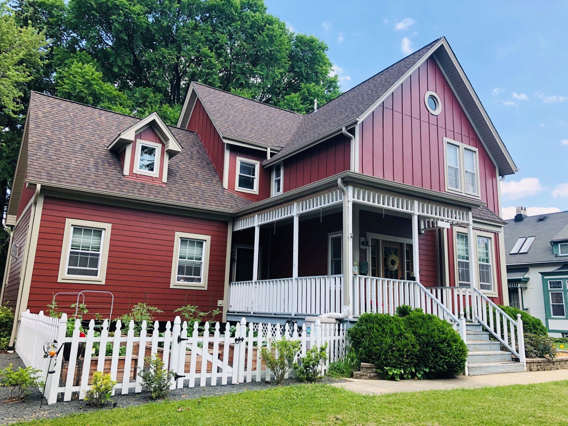 A charming red house with white trim and a front porch, surrounded by a white picket fence and greenery under a clear blue sky.
