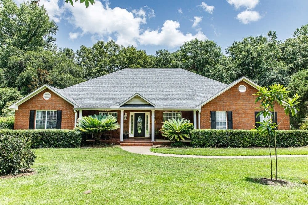 A red brick ranch-style home with a gray shingle roof, white trim, and a covered front porch, set against a backdrop of lush green trees and a bright blue sky.