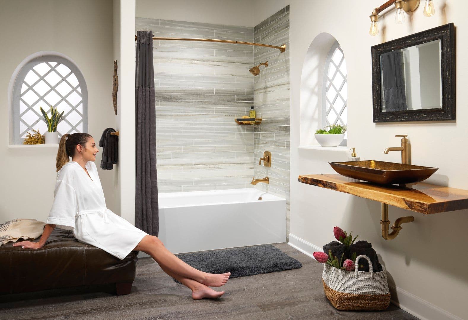Woman in white robe sitting on leather bench in luxury bathroom with soaking tub, vessel sink, and decorative arched windows