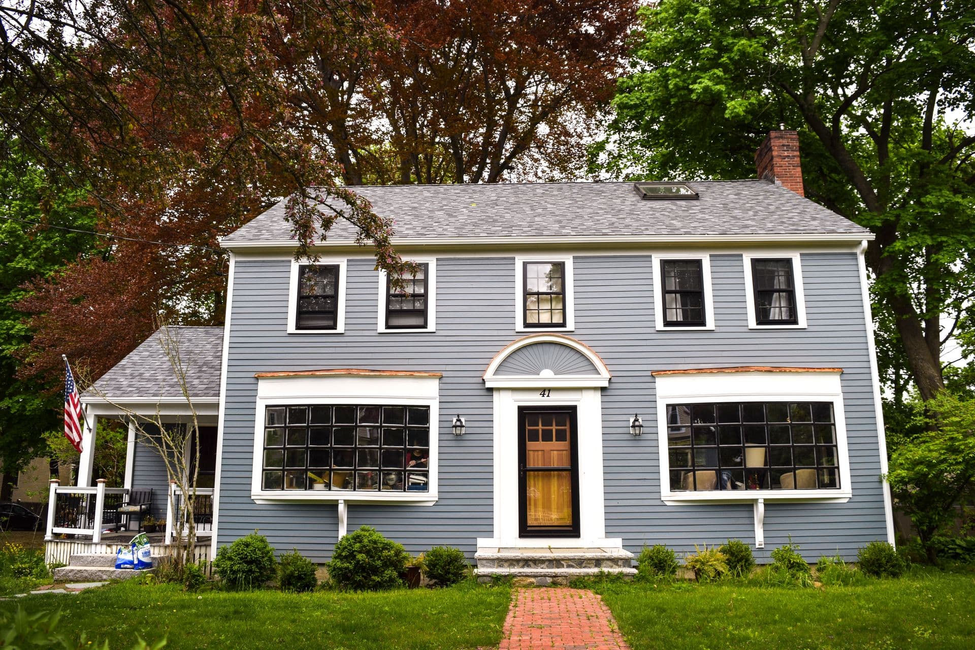 A two-story blue colonial-style house with white trim, a brick walkway, and surrounded by trees and greenery.