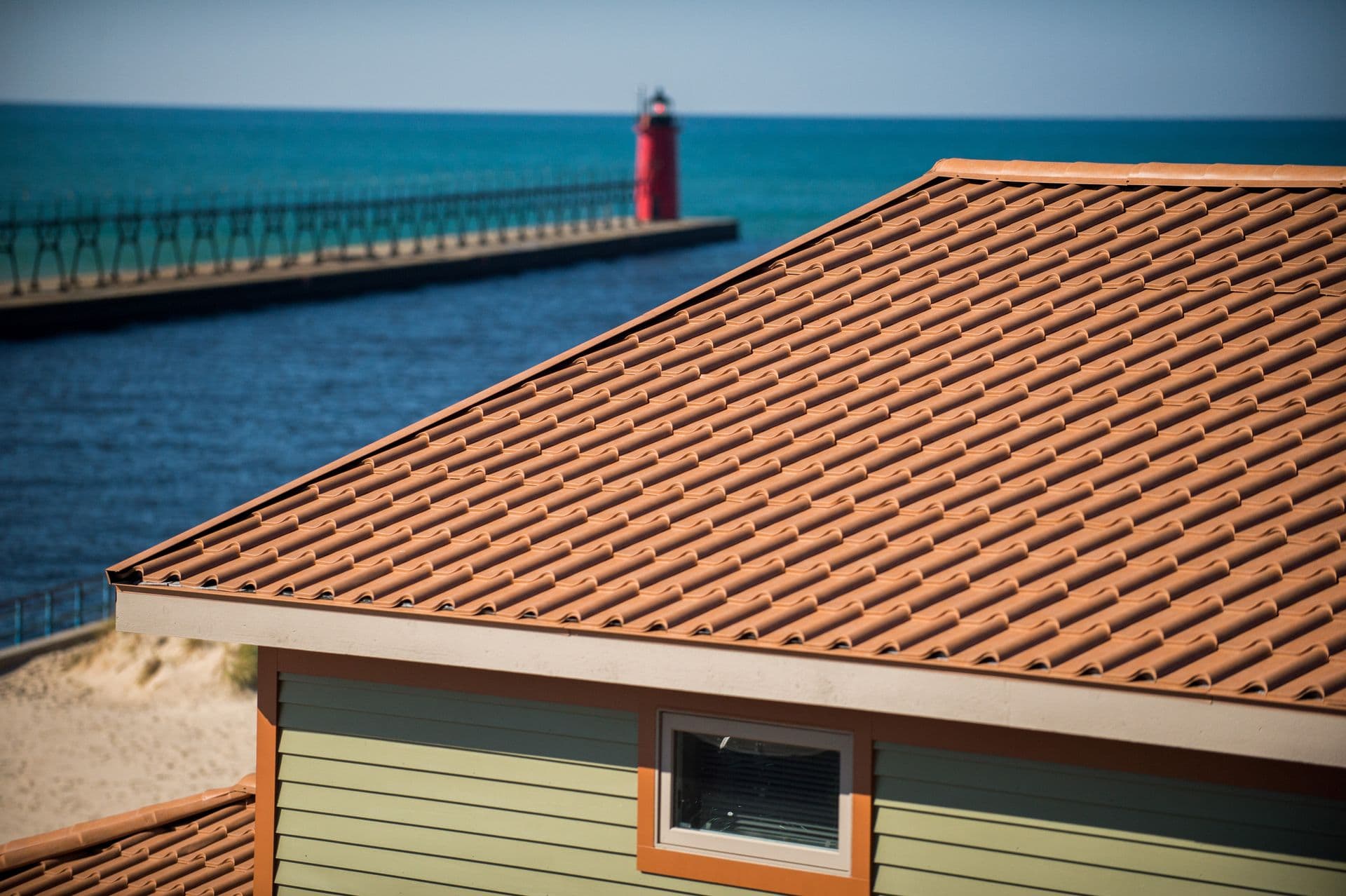 A close-up of a terracotta-tiled roof on a coastal house with a red lighthouse in the background.