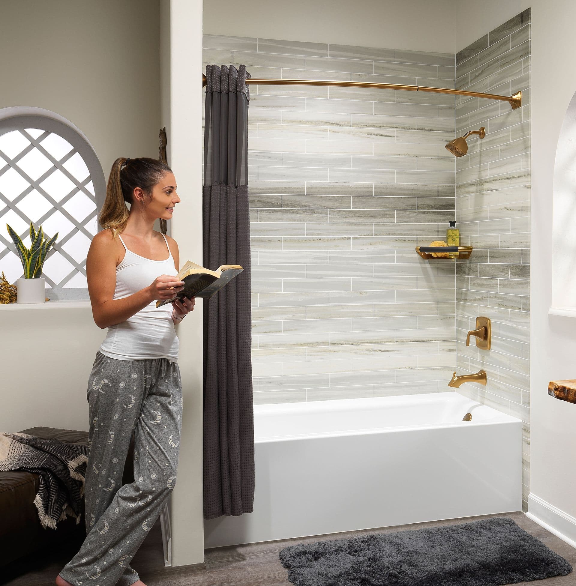 A woman in pajamas reads a book beside a modern bathtub with marble tiles and gold fixtures.