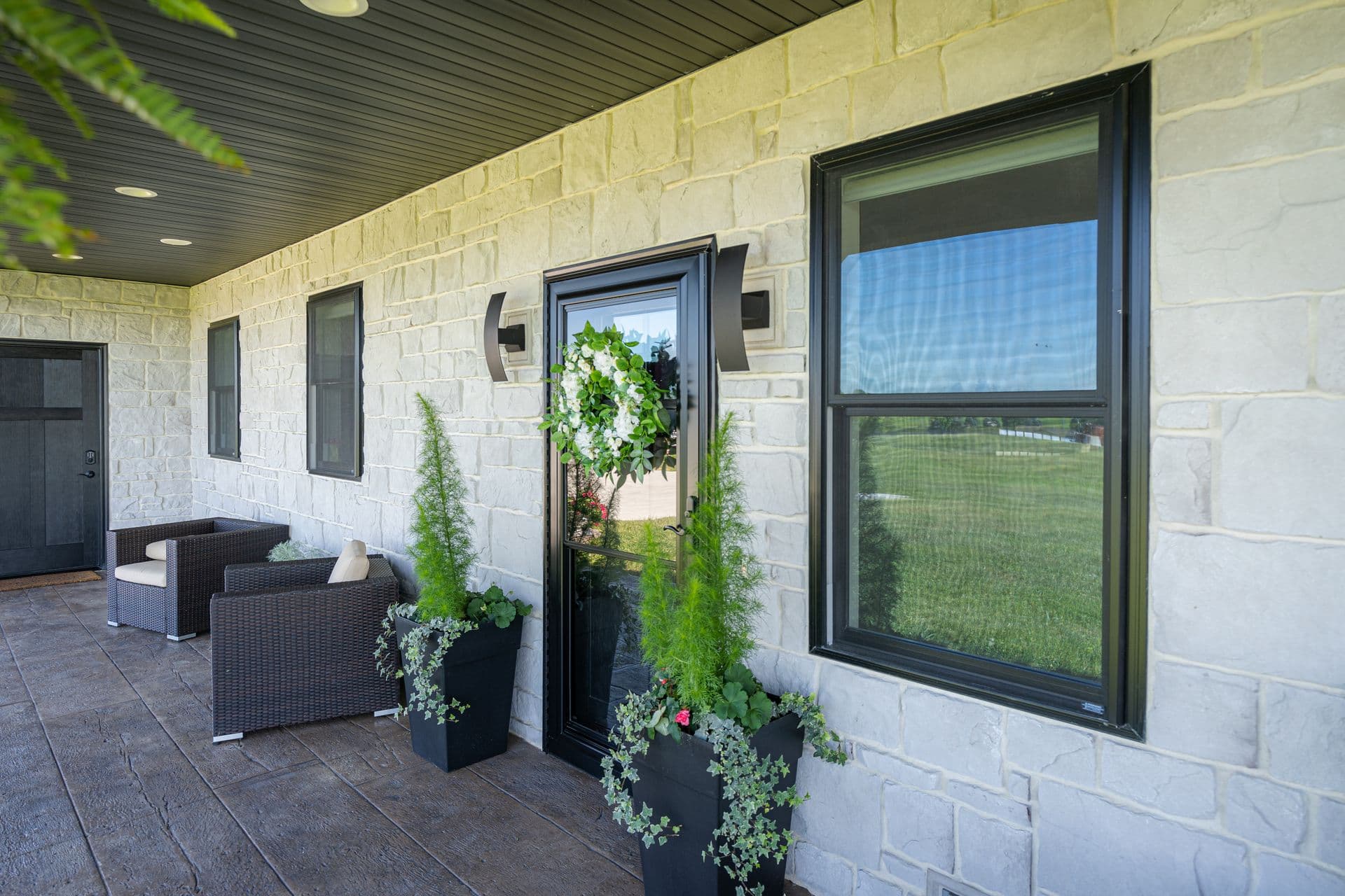 A covered stone porch features black-framed windows, a glass door adorned with a floral wreath, and dark wicker seating flanked by tall potted plants.