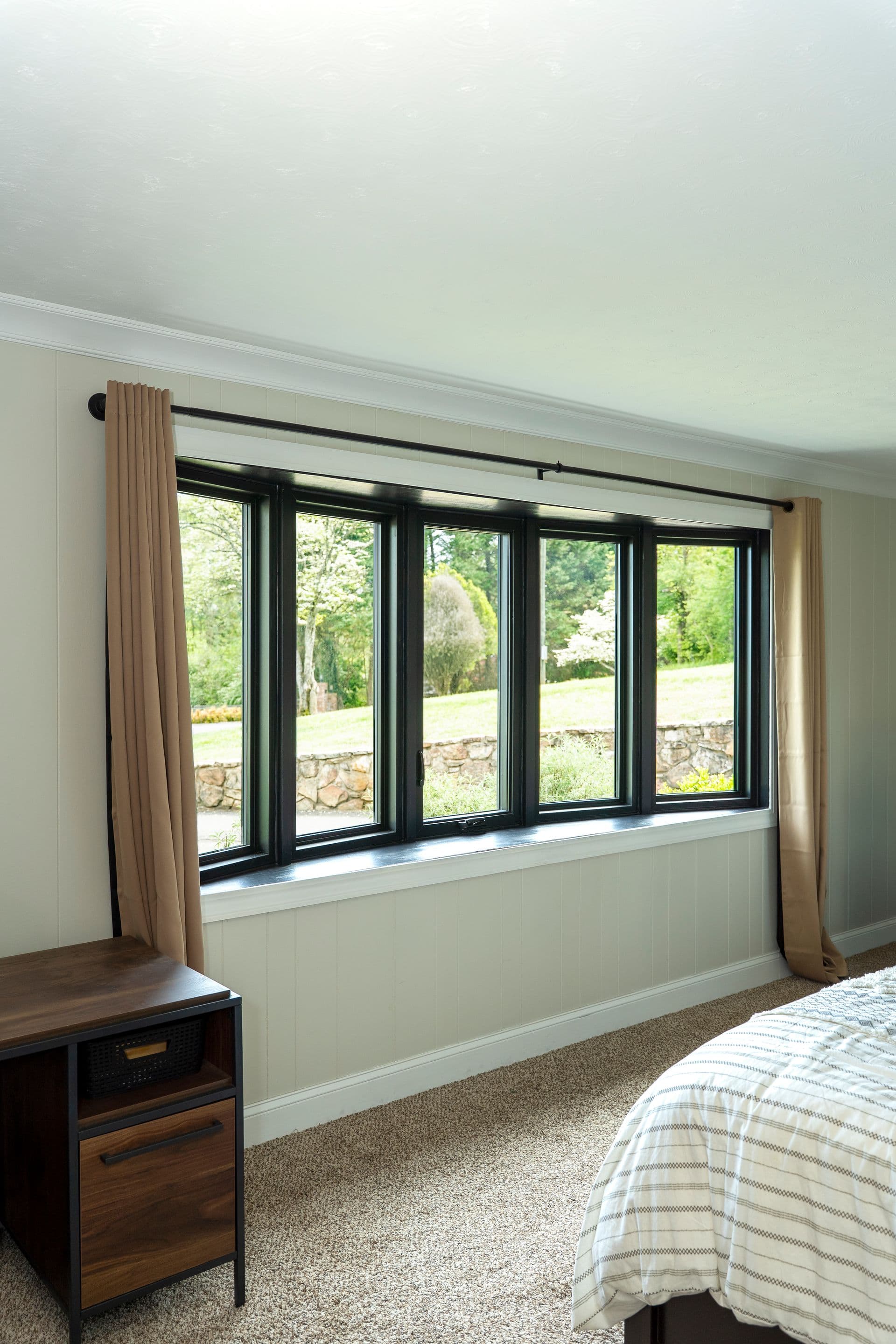 A serene bedroom features a large black-framed window with beige curtains, offering a view of a lush green yard and stone wall.