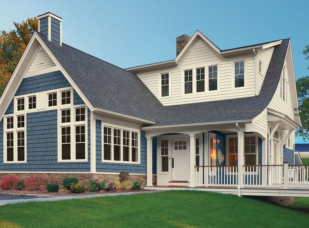 A charming two-story house with blue shingle siding and white trim, featuring a covered porch and large windows under a dark roof.