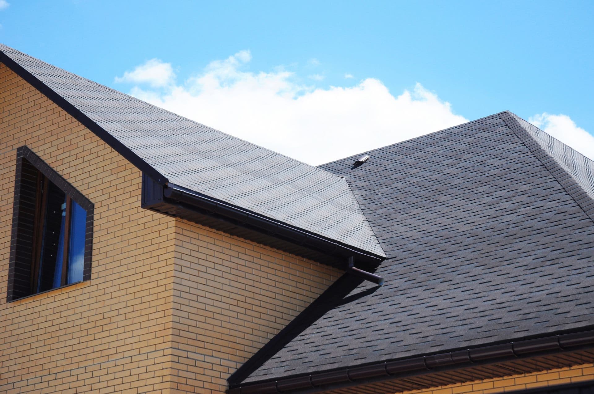 A close-up of a modern house's dark shingled roof against a bright blue sky with scattered clouds.