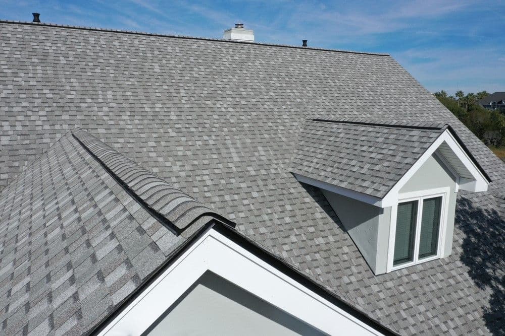 Aerial view of a gray shingle roof with white trim and a dormer window under a blue sky.
