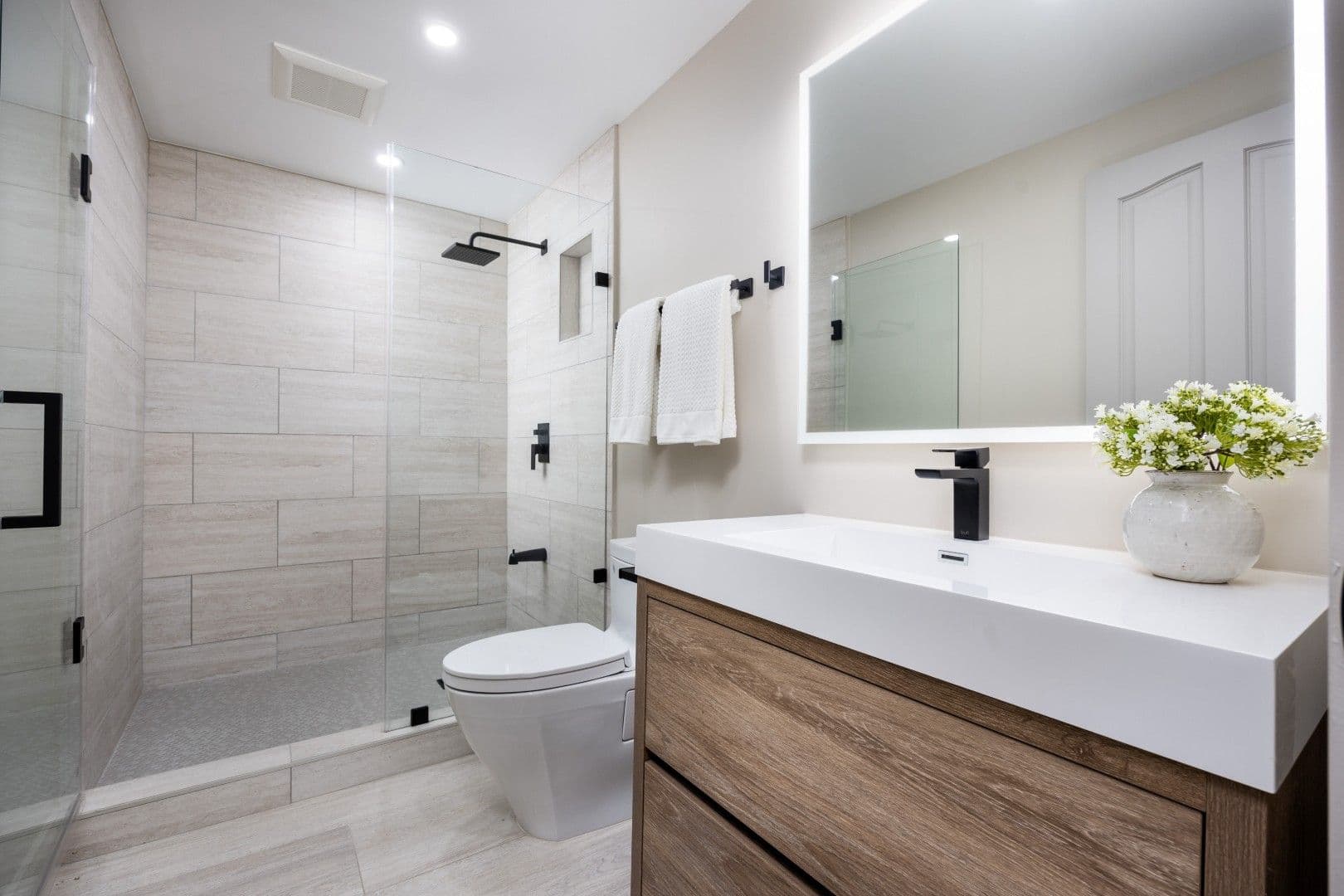 A sleek, contemporary bathroom featuring a glass-enclosed shower, wooden vanity, and black fixtures against neutral tones.