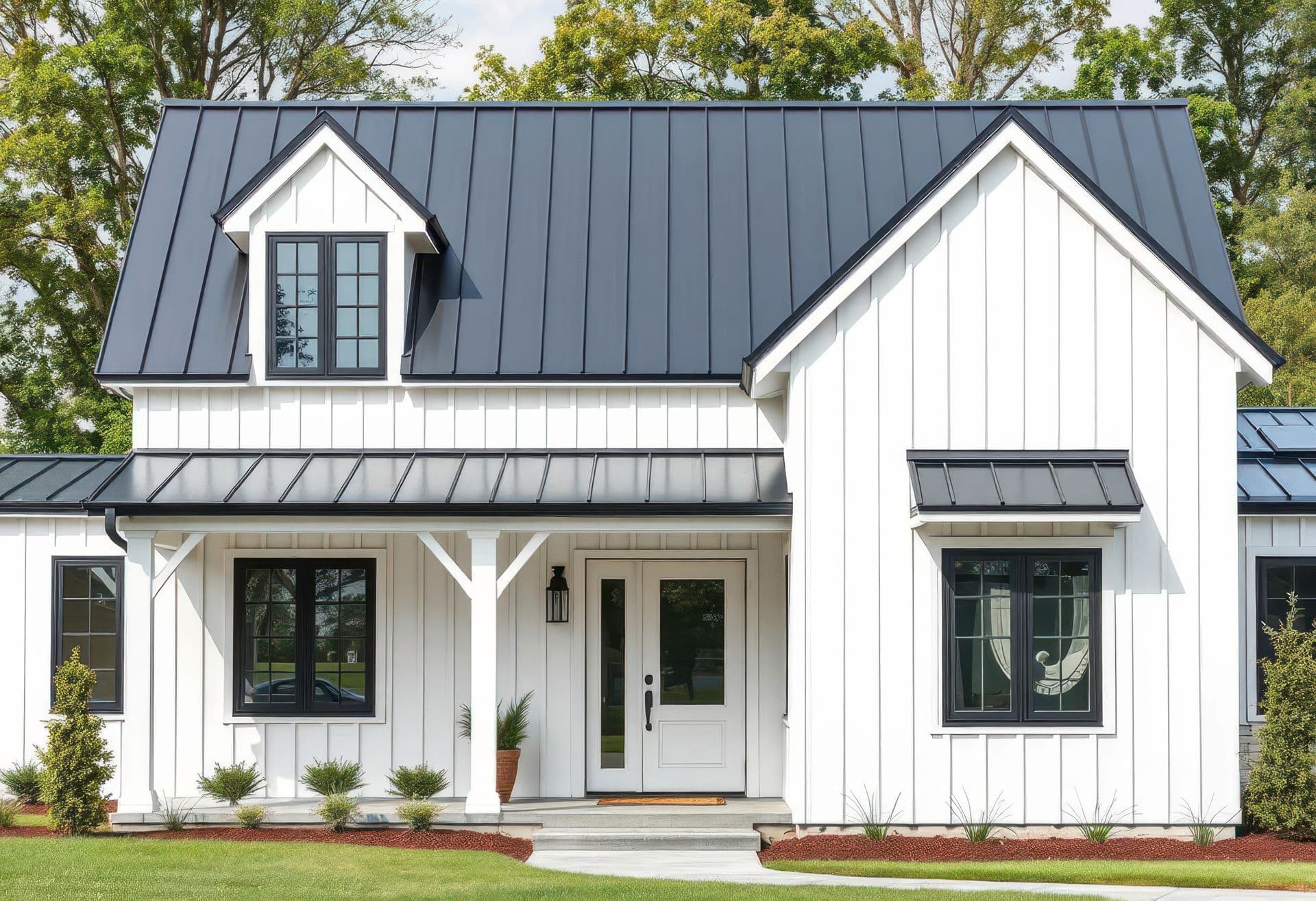 A white clapboard house with a dark gray metal roof and black trim features a covered porch and lush green landscaping.
