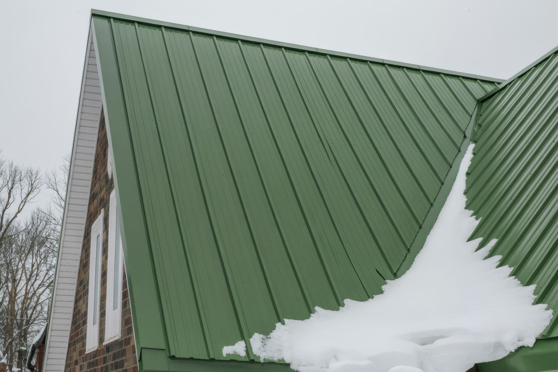A close-up of a green corrugated metal roof with snow accumulation on its edge against a gray sky.