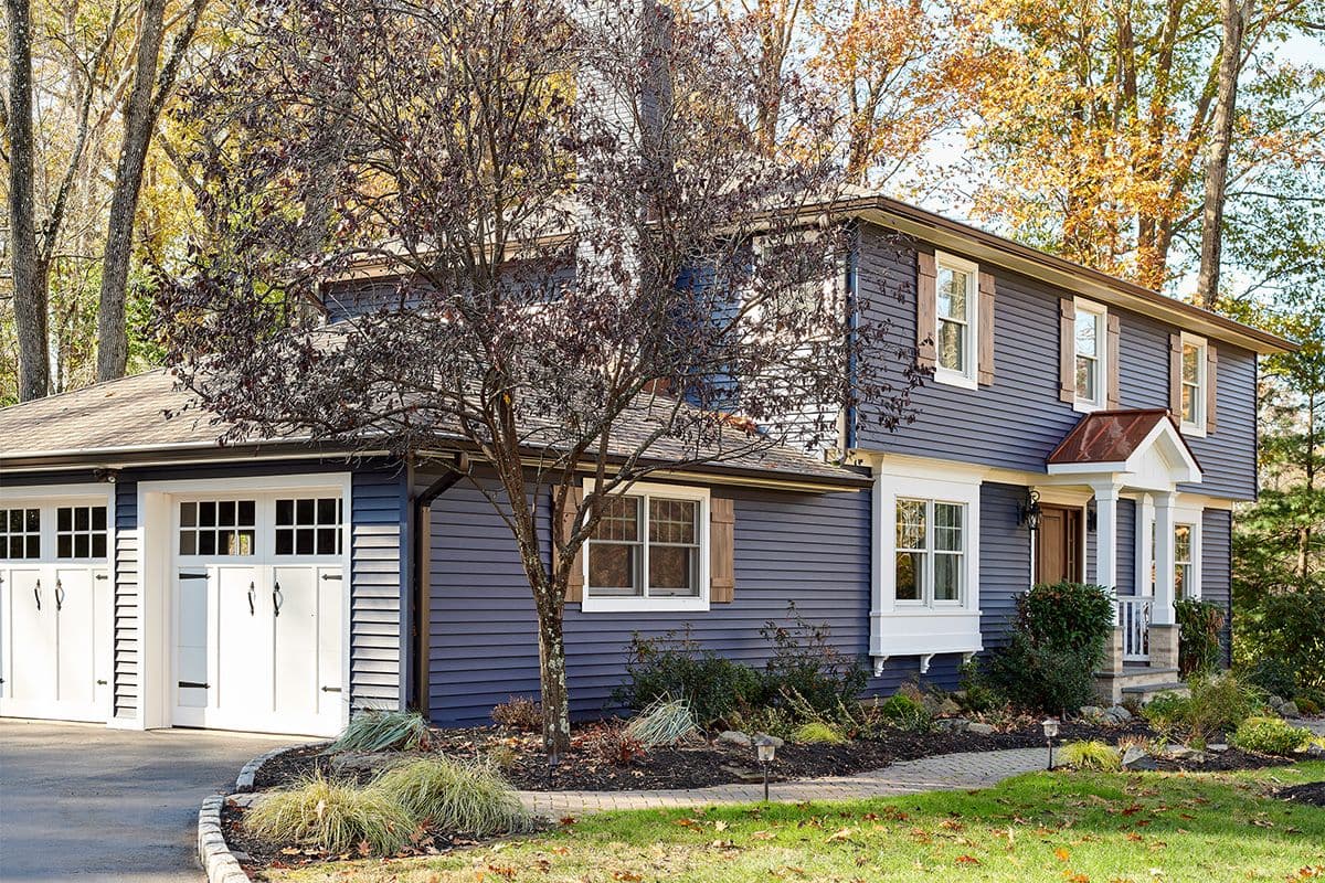 A two-story blue house with white trim and a garage, surrounded by autumn trees and landscaped yard.